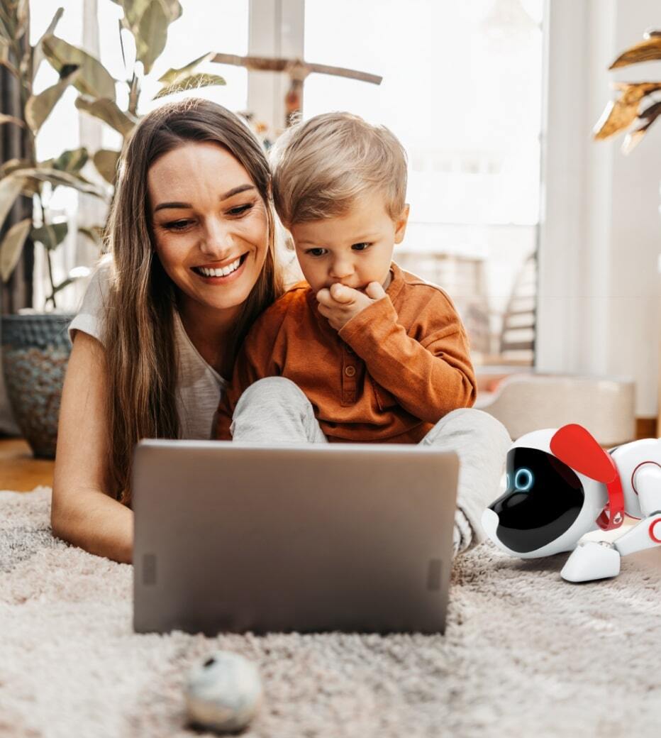 Mãe e filho a olhar para o computador e a sorrir, com o Mealheiro Inteligente Trusty ao seu lado